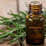 Frasco de vidro âmbar com óleo de alecrim artesanal sobre mesa rústica de madeira, ao lado de ramos frescos de alecrim